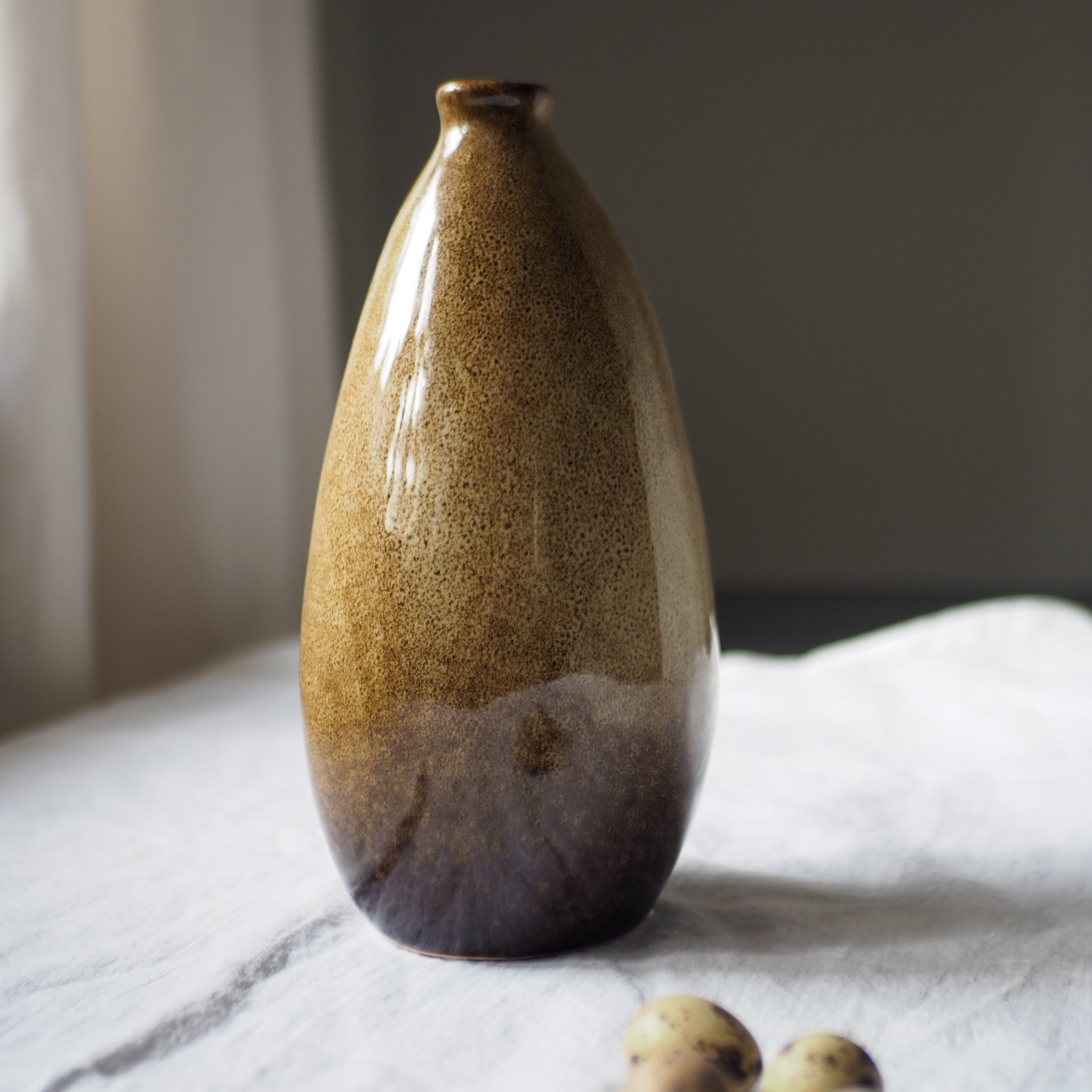 Dwa małe, ręcznie robione wazony porcelanowe w stylu japandi. Wazony mają wysokość 23 cm i pękaty kształt, dostępne w wariancie kremowym nakrapianym oraz brązowym cieniowanym. Fotografia na białym obrusie z naturalnymi dodatkami.